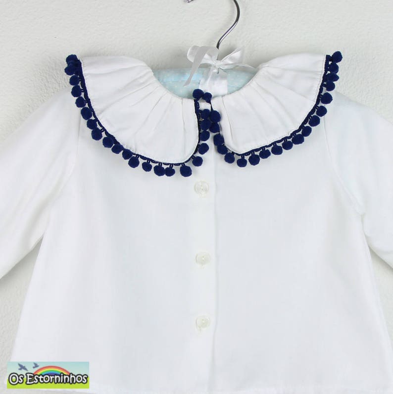 Op de afbeelding: Een wit overhemd met knopen en een grote, gerimpelde kraag afgezet met marineblauwe pompons. Het shirt heeft lange mouwen en hangt aan een zilveren hanger met een wit lint. De tekst "Os Estorninhos" staat in de linker benedenhoek.