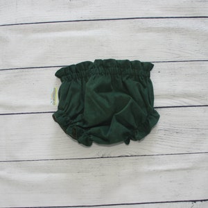 May include: Dark green baby bloomers with a ruffled waistband and leg openings. The bloomers are made of a soft, textured fabric and are displayed on a white wooden surface.