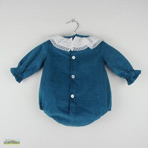 May include: Teal-colored baby romper with long sleeves and a white lace collar. The romper has three white buttons down the front and ruffled cuffs. The garment is hanging on a silver hanger against a white background.