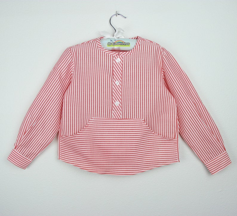 May include: A red and white striped long-sleeved shirt with a mandarin collar and three white buttons. The shirt has a unique pocket design and is hanging on a metal hanger.