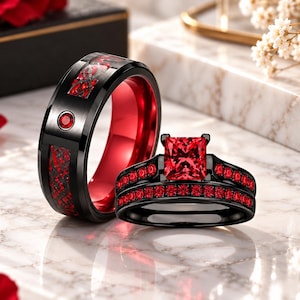 Puede incluir: Un conjunto de dos anillos que incluye una banda negra con interior rojo e incrustaciones de fibra de carbono roja, y un anillo de compromiso negro con una piedra central roja de corte princesa y banda pavé con pequeñas piedras rojas.