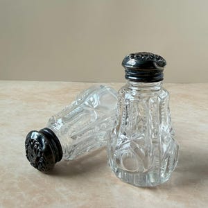 May include: Two clear glass shakers featuring a faceted, geometric cut-glass pattern and rounded bodies. Each shaker is topped with a dark, tarnished metal cap that displays an intricate, embossed floral or scrollwork design on the top surface.