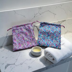 Puede incluir: Dos bolsas florales con cordón y cordones morados, una en rosa y morado, la otra en azul y morado. Un pequeño cuenco blanco con líquido amarillo y una toalla blanca enrollada también están presentes sobre una superficie de mármol.