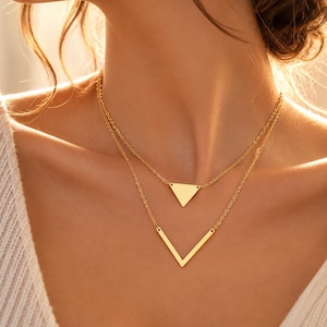 Puede incluir: Un collar de capas en tono dorado con dos cadenas delicadas. La cadena superior sostiene un pequeño dije triangular sólido, mientras que la cadena inferior, más larga, presenta un dije en forma de V. Ambos dijes geométricos tienen un acabado liso.