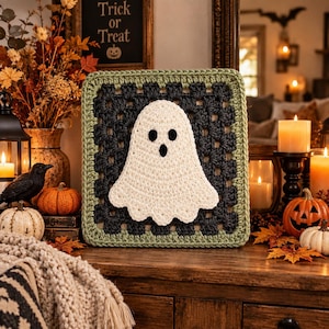 May include: A square crochet granny square featuring a central white ghost applique with black eyes and a mouth. The square has a dark charcoal gray center and a sage green border. The ghost is centered on the dark gray crochet background.