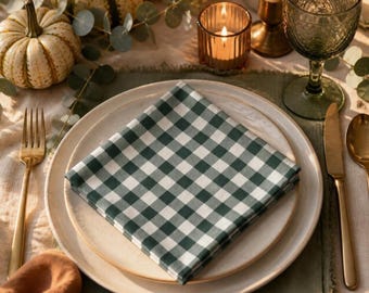 May include: A square cloth napkin featuring a dark green and white gingham check pattern, folded and placed centrally on a round off-white ceramic dinner plate. The napkin is positioned on a table setting with a gold fork, knife, and spoon.