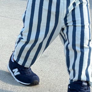 May include: A pair of white trousers with vertical navy blue stripes paired with a yellow, white, and blue horizontal striped shirt and navy blue athletic trainers featuring a white N logo and the number 515 on the tongue of the shoe.