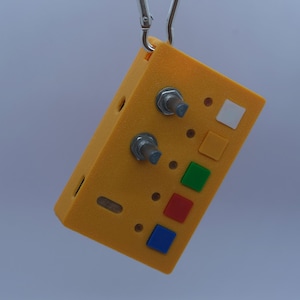 May include: A rectangular yellow electronic device with a textured finish, suspended by a silver metal carabiner. The front panel features two metallic rotary knobs and four square buttons in white, yellow, green, red, and blue colors.