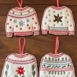 May include: Four white felt Christmas sweater ornaments with red and white striped hanging ribbons. Each ornament features a different embroidered design, including snowflakes, stars, and floral patterns, with red and blue accents.