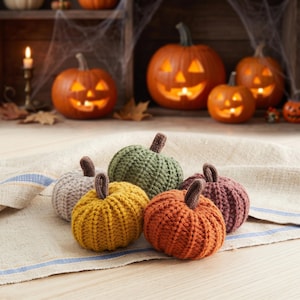 May include: A collection of six small, round, crocheted pumpkins in various colors including beige, mustard yellow, olive green, orange, and dark red, each featuring a small brown stem on top, resting on a textured off-white fabric.