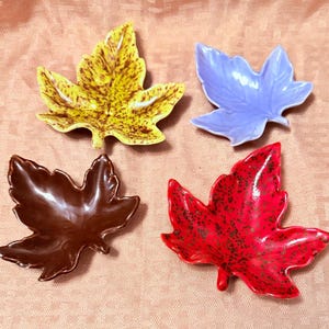 May include: A set of four ceramic leaf-shaped dishes. One dish is yellow with brown speckles, one is solid light purple, one is solid dark brown, and one is bright red with dark green speckles. Each dish features a detailed veined texture.