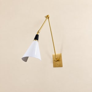 Può includere: Una lampada da parete con finitura in ottone, piastra rettangolare e braccio orientabile regolabile. La lampada presenta un paralume conico con esterno bianco e un dettaglio nero sulla parte superiore, offrendo una luce direzionata.