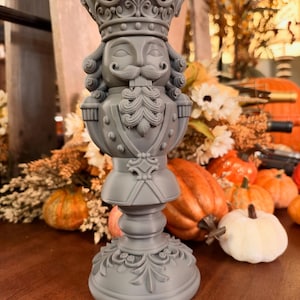 May include: A matte gray nutcracker figurine featuring a detailed crown, a curled mustache, and a beard. The figure stands on a circular base with ornate scrollwork patterns and has a uniform, solid-colored finish throughout its structure.