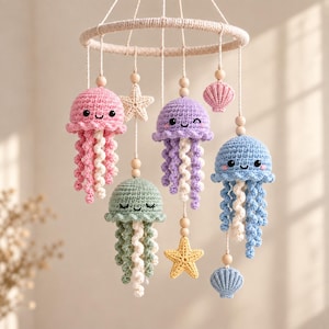 May include: A hanging mobile featuring four crocheted jellyfish in pink, purple, light blue, and sage green, each with curly tentacles. The mobile includes two crocheted starfish, two crocheted seashells, and wooden beads suspended from a circular frame.