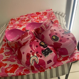 May include: A pair of pink camouflage clogs featuring a bright pink interior and adjustable heel straps. The shoes are decorated with various rubber charms, including a shark mouth, a monkey face, and text that reads APE NEVER APE.