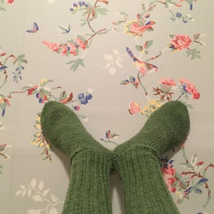 Puede incluir: Un par de calcetines verdes de punto sobre un fondo de papel pintado floral. Los calcetines son de color verde liso y parecen hechos a mano. El papel pintado presenta un estampado de flores rosas, amarillas y azules con mariposas.
