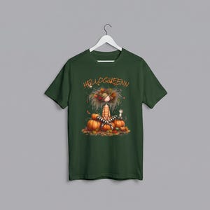 Peut inclure: Un t-shirt vert foncé à manches courtes avec une impression graphique représentant un personnage aux cheveux ébouriffés assis parmi des citrouilles orange et des feuilles d'automne. Le texte HELLOQUEENN est imprimé en orange sur le devant.