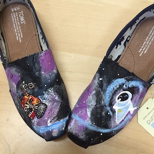 May include: A pair of black canvas shoes with a galaxy print. One shoe features a painted image of Wall-E, the other features a painted image of EVE. The shoes have a brown insole with the TOMS logo and the text "With every pair you purchase, TOMS will give a pair of new shoes to a child in need. One for One."
