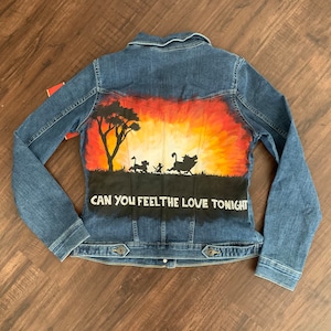 May include: A blue denim jacket with a hand-painted design featuring a sunset scene with silhouettes of characters and the text "CAN YOU FEEL THE LOVE TONIGHT". The jacket has long sleeves and a button-up front.