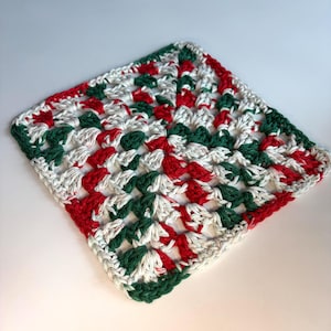 May include: A square crochet coaster or dishcloth featuring a classic granny square pattern. The textile is crafted with alternating yarn colors in vibrant red, forest green, and off-white, creating a repeating geometric design.