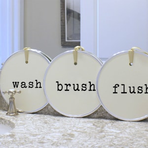 Op de afbeelding: Drie ronde, witte decoratieve borden met zwarte letters. De borden lezen "wash", "brush" en "flush".