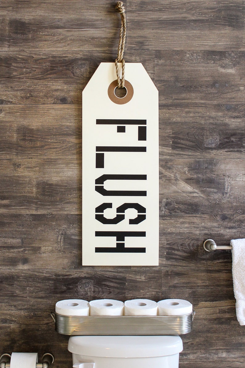 以下が含まれることがあります： 茶色の木製の壁に「FLUSH」と黒文字で書かれた白い看板が掛かっています。白いトイレには銀色のトイレットペーパーホルダーとトイレットペーパーのロールが1つ付いています。銀色のラックにはトイレットペーパーのロールが5つあります。