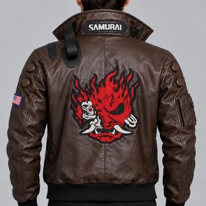 Puede incluir: Una chaqueta bomber de cuero marrón con un gráfico grande de una máscara oni roja y negra en la espalda. El cuello muestra el texto SAMURAI. La chaqueta incluye un pequeño parche de la bandera estadounidense en la manga derecha y puños acanalados negros.