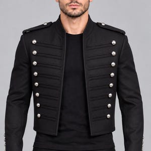 May include: A black military-style jacket featuring a high collar, long sleeves, and shoulder epaulets. The front is adorned with two vertical rows of silver-toned buttons and horizontal cord detailing, worn over a plain black t-shirt and black pants.