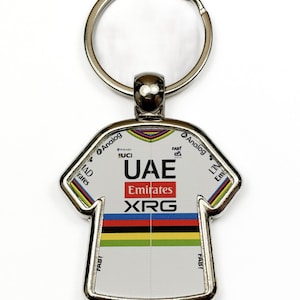 May include: A metal keyring shaped like a cycling jersey. The white jersey features the text UAE, Emirates, and XRG, along with UCI and Analog logos. A horizontal band of blue, red, black, yellow, and green stripes runs across the centre.