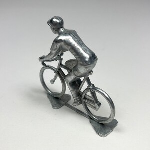 May include: A silver metal figurine of a cyclist riding a bicycle. The cyclist is wearing a cap and a short-sleeved shirt. The bicycle has a silver frame and wheels.