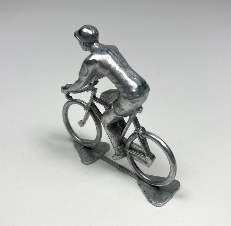 May include: A silver metal figurine of a cyclist riding a bicycle. The cyclist is wearing a cap and a short-sleeved shirt. The bicycle has a silver frame and wheels.