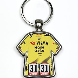 May include: A yellow jersey-shaped keychain with the Visma Lease a Bike logo and the numbers 31 31. The keychain has a silver metal finish and a ring for attaching keys. The jersey design includes sponsor logos.