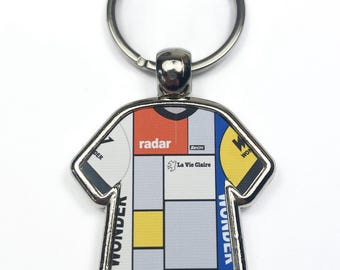 Può includere: Un portachiavi in metallo argentato a forma di maglia da ciclismo con un motivo geometrico bianco, rosso, giallo e blu. La maglia ha il testo "radar" e "La Vie Claire" stampato su di essa.