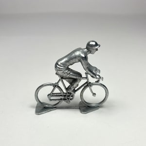 May include: A silver metal figurine of a cyclist riding a bicycle. The figurine is detailed with a helmet, jersey, shorts, and shoes. The bicycle has two wheels, handlebars, and a seat.