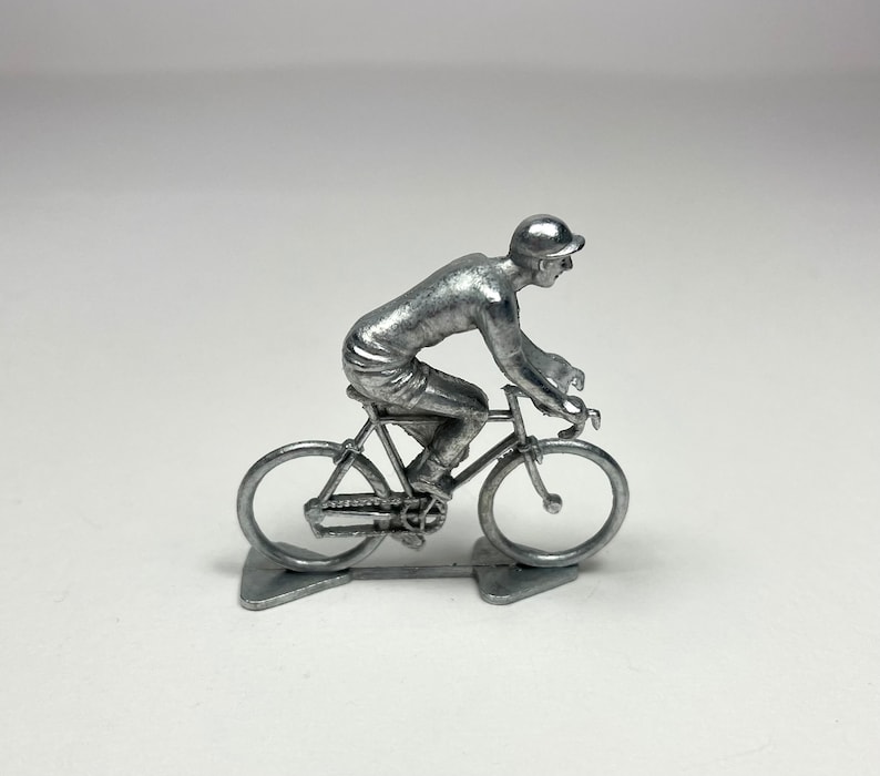 May include: A silver metal figurine of a cyclist riding a bicycle. The figurine is detailed with a helmet, jersey, shorts, and shoes. The bicycle has two wheels, handlebars, and a seat.