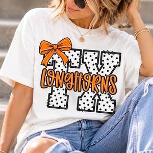 Może przedstawiać: Biały t-shirt z krótkim rękawem z dużym nadrukiem graficznym przedstawiającym litery TX w kolorze białym w czarne kropki z czarnymi przeszyciami. Na literach znajduje się pomarańczowa kokarda, a słowo LONGHORNS jest napisane pomarańczową czcionką.