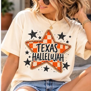 Könnte beinhalten: Ein cremefarbenes Kurzarm-T-Shirt mit einem großen, zentralen, orange-weiß karierten Sternmotiv mit texturiertem Rand. Der Schriftzug "TEXAS HALLELUJAH" ist in Schwarz über den Stern gedruckt und von mehreren schwarzen, gesprenkelten Sternen umgeben.