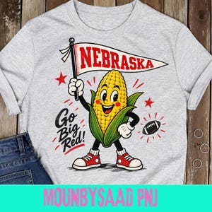 Może przedstawiać: Jasnoszary t-shirt z rysunkową postacią kolby kukurydzy w czerwonych trampkach, trzymającą biały proporczyk z czerwonym napisem NEBRASKA. Grafika zawiera czarny napis Go Big Red!, czerwone gwiazdki oraz czarno-białą piłkę do futbolu.