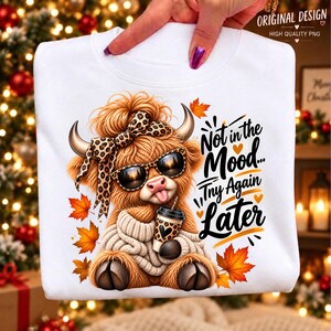 Puede incluir: Una sudadera blanca con el gráfico de una vaca Highland marrón y peluda con un lazo y gafas de sol de leopardo, sosteniendo un café. El texto "Not in the Mood... Try Again Later" está rodeado de hojas de otoño naranjas y amarillas.