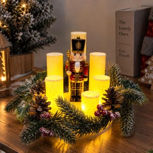Pode incluir: Um centro de mesa de Natal com uma figura central de quebra-nozes rodeada por cinco velas cilíndricas iluminadas. O arranjo está inserido numa coroa de ramos de pinheiro verdes, pinhas castanhas e pequenas bagas vermelhas com detalhes de neve.
