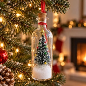 May include: A clear glass bottle ornament containing a miniature green Christmas tree with colorful baubles, topped with a gold star, and set in white artificial snow. The bottle is sealed with a cork and hangs from a red ribbon.