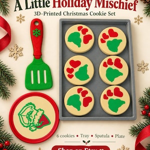 Pode incluir: Um conjunto de biscoitos de Natal impressos em 3D com seis biscoitos redondos com corações vermelhos e pegadas verdes numa bandeja cinzenta, uma espátula verde e vermelha e um prato redondo com um gráfico de personagem. Texto A Little Holiday Mischief.