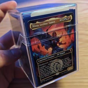 Peut inclure: Une boîte de jeu en plastique transparent contenant une carte à collectionner Magic: The Gathering intitulée Nissa, Leyline Tamer. La carte présente un personnage elfe magicien légendaire avec des illustrations orange et bleues.