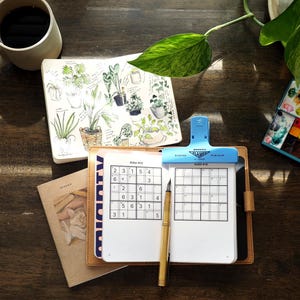 May include: A tan leather notebook cover holding a Sudoku puzzle book with a blue plastic clip at the top, a gold fountain pen, a sketchbook with botanical illustrations, a ceramic mug with dark liquid, and a small watercolour paint set.