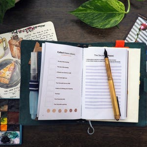 May include: A dark teal leather journal open to a page titled Collect these colours and The Secret Poem, featuring a gold fountain pen, a water brush, a watercolor palette, a sketchbook with illustrations, and two small photographs.