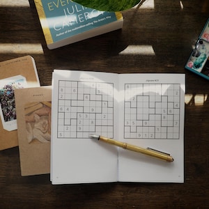 Könnte beinhalten: Ein offenes Rätselbuch mit zwei Sudoku-Gittern, beschriftet als Jigsaw #30 und Jigsaw #31, mit einem goldfarbenen Füllfederhalter, der diagonal über den Seiten liegt. Zwei weitere Notizbücher mit illustrierten Einbänden und ein Aquarellkasten liegen daneben.