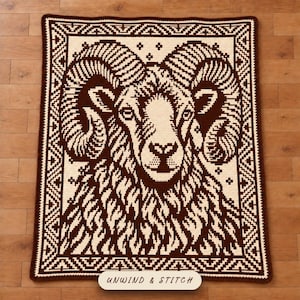 May include: A rectangular crochet blanket featuring a dark brown and cream color scheme. The design displays a large ram head with curved horns at the center, surrounded by a geometric border. The text UNWIND & STITCH is printed at the bottom.