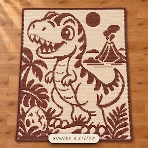 May include: A rectangular crochet blanket featuring a two-tone brown and cream design of a cartoon dinosaur, a volcano, palm leaves, and an egg. The text "UNWIND & STITCH" is printed on a label at the bottom centre of the textured fabric piece.
