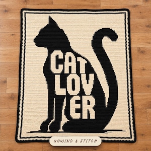 Peut inclure: Une couverture en crochet rectangulaire présentant une grande silhouette noire de chat assis sur un fond crème. Le texte CAT LOVER est intégré dans le corps du chat en couleur crème. La couverture possède une épaisse bordure noire.