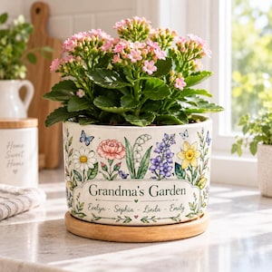 Könnte beinhalten: Ein weißer Keramikübertopf mit einer bunten Blumenillustration, dem Text "Grandma's Garden" und den Namen "Evelyn - Sophia - Linda - Emily". Der Übertopf enthält eine rosa Kalanchoe und steht auf einem runden Holzsockel.
